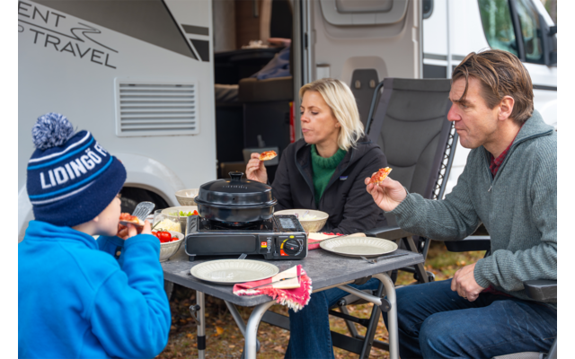 Frau, Mann und Junge sitzen an einem Campingtisch vor einem Wohnmobil; auf dem Tisch stehen ein tragbarer Gaskocher, Teller, eine Schüssel und Brotstücke; der Junge trägt eine blaue Mütze, die Frau einen grünen Schal