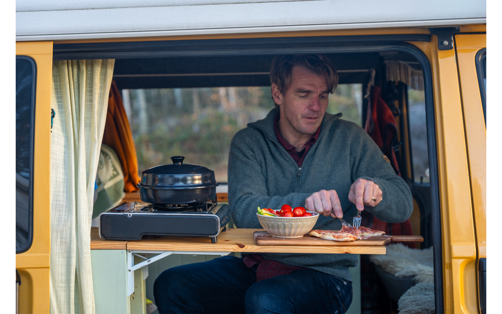 Mann sitzt in offenem gelben Campervan und schneidet auf einem Brett; auf dem Tisch ein tragbarer Kocher mit Topf und eine Schüssel Kirschtomaten