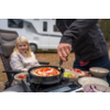 Person dreht Pfanne mit Pizza auf tragbarem Gaskocher; Schalen mit Salat und geschnittenen Zwiebeln auf dem Tisch; blondes Mädchen sitzt auf Campingstuhl vor Wohnmobil