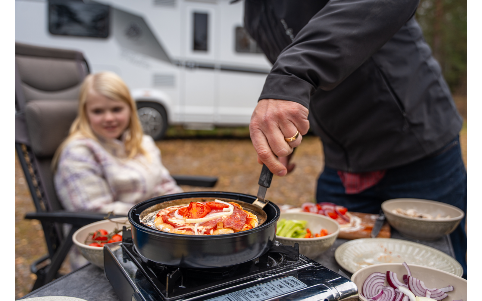 Person dreht Pfanne mit Pizza auf tragbarem Gaskocher; Schalen mit Salat und geschnittenen Zwiebeln auf dem Tisch; blondes Mädchen sitzt auf Campingstuhl vor Wohnmobil
