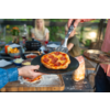 Kleine Pepperoni-Pizza auf schwarzem Teller, mit Spachtel serviert; im Hintergrund Tisch mit Schalen, Mehl, weiterer Pizza und verschwommenen Personen