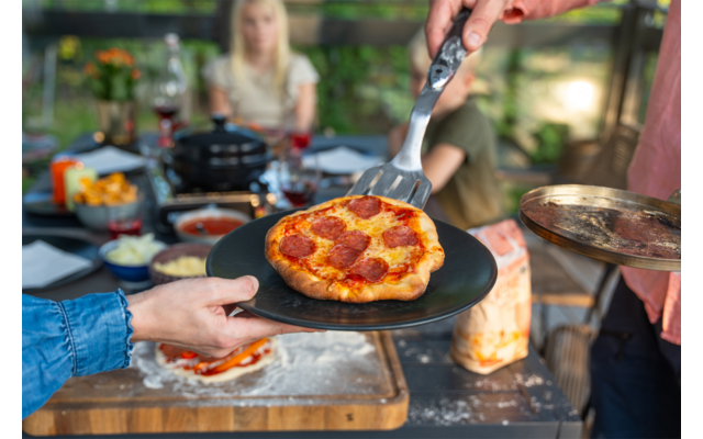 Kleine Pepperoni-Pizza auf schwarzem Teller, mit Spachtel serviert; im Hintergrund Tisch mit Schalen, Mehl, weiterer Pizza und verschwommenen Personen