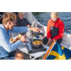 Eine erwachsene Person kocht Gemüse in einer Pfanne auf einem mobilen Herd an Bord eines Bootes; zwei Kinder sitzen daneben, Wasser im Hintergrund, schwedische Flagge sichtbar