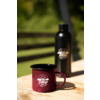 Tasse émaillée bordeaux avec logo blanc «camp life» sur table en bois ; gourde noire avec logo en arrière-plan, fond vert flou et lattes de bois horizontales