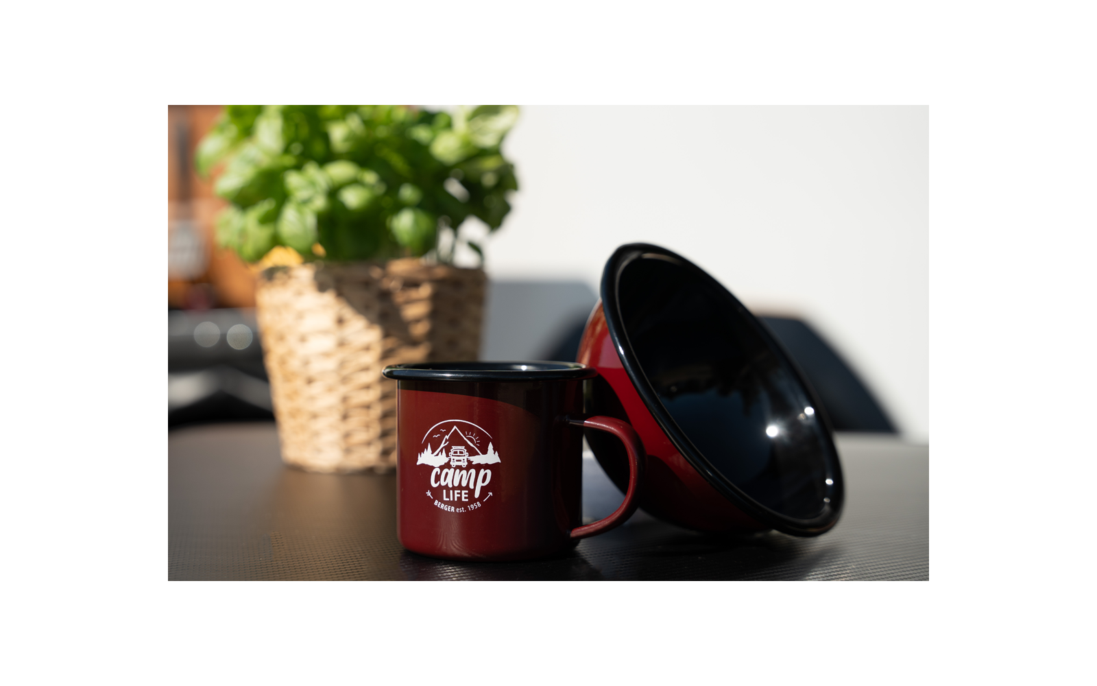 Tasse de camping en émail rouge avec logo blanc à côté d'une tasse retournée, plante de basilic dans un panier tressé en arrière-plan sur une table noire