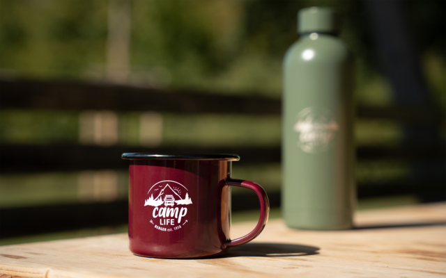 Roter Emaille-Campingbecher mit weißem "camp life"-Logo vorn auf einem Holztisch; unscharfe grüne Thermosflasche im Hintergrund