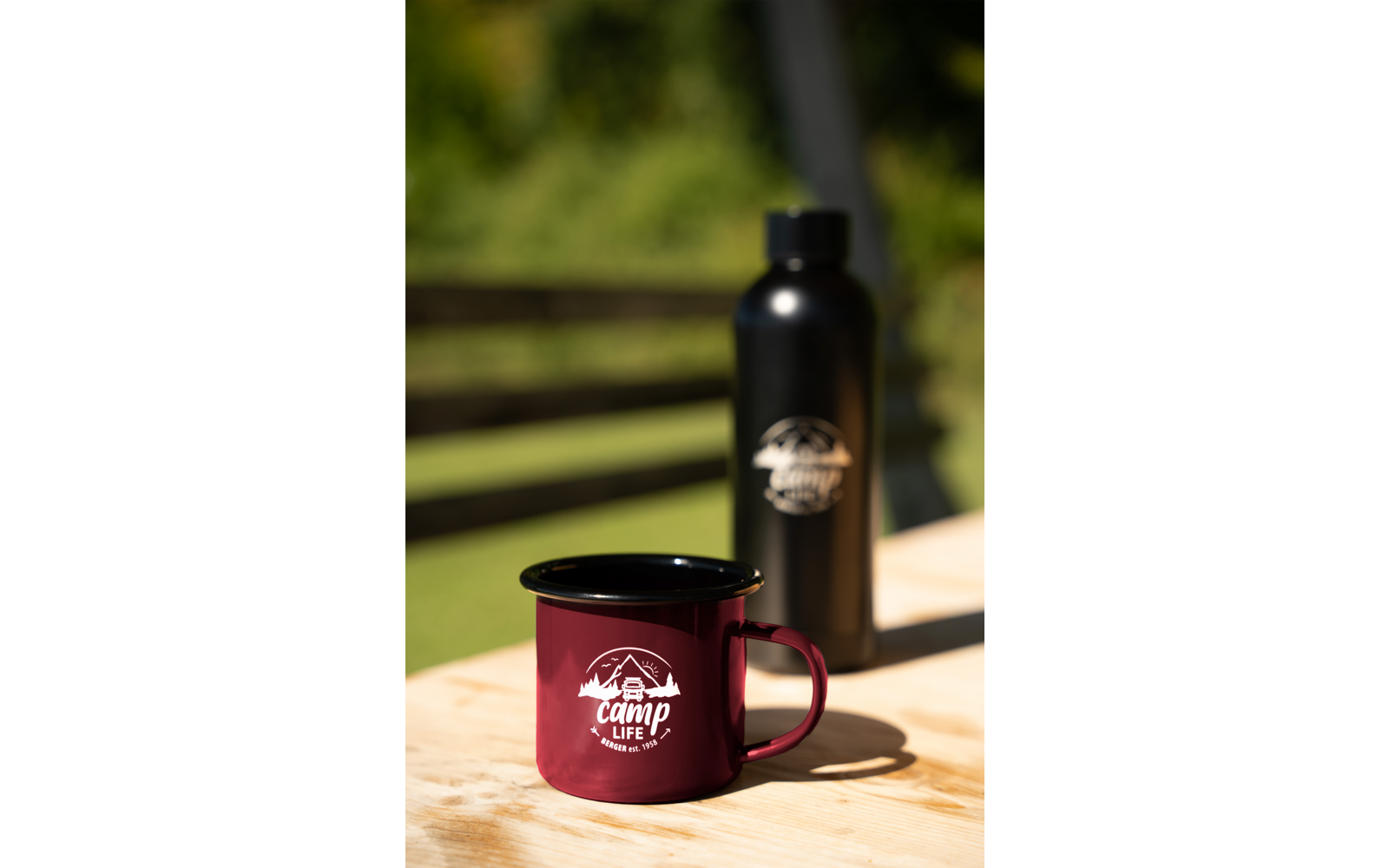Bordeauxrote Emaille-Tasse mit weißem "camp life"-Logo auf Holztisch; im Hintergrund schwarze Trinkflasche mit Logo, unscharfer grüner Hintergrund und horizontale Holzlatten