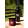 Bordeauxrote Emaille-Tasse mit weißem "camp life"-Logo auf Holztisch; im Hintergrund schwarze Trinkflasche mit Logo, unscharfer grüner Hintergrund und horizontale Holzlatten