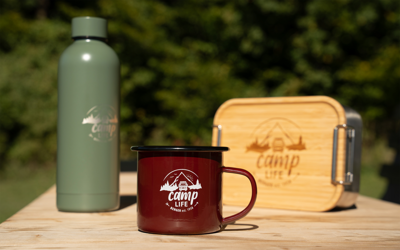 Grüne Thermosflasche, roter Emaille-Becher mit Aufschrift 'camp life' und Holzbox mit 'camp life'-Logo auf einem Holztisch vor unscharfem Waldhintergrund