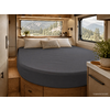 Interieur van een camper met een rond bed met donkergrijs hoeslaken, drie kussens (twee wit, één beige), houten kasten en lades, ramen met zicht op dennenbomen en bergen, een potplant op het aanrecht
