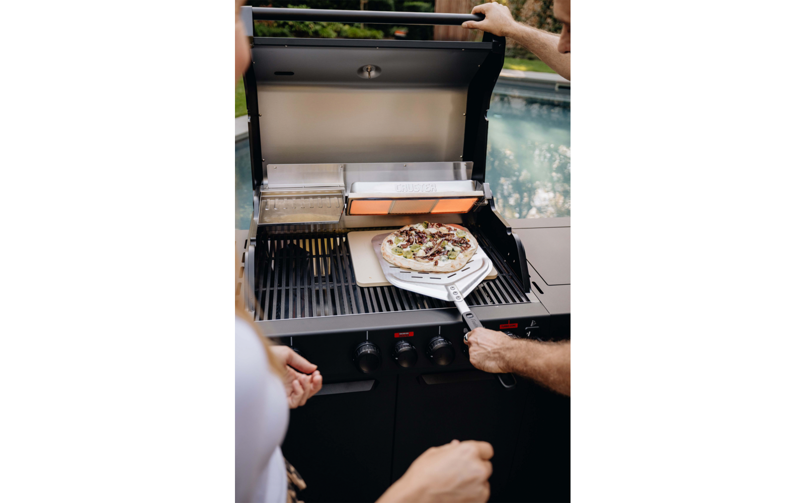 Offener Gasgrill mit Pizza auf einem Pizzaschieber, zwei Personen nur mit Armen sichtbar, Swimmingpool im Hintergrund