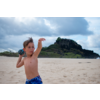 Junge mit blauem Badeanzug am Strand, im Hintergrund Felsen und bewölkter Himmel