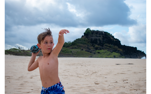 Junge mit blauem Badeanzug am Strand, im Hintergrund Felsen und bewölkter Himmel