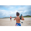 Junge mit blauer Badehose und Wasserpistole, Strand im Hintergrund, andere Person entfernt sichtbar