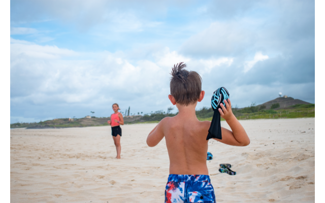 Junge mit blauer Badehose und Wasserpistole, Strand im Hintergrund, andere Person entfernt sichtbar