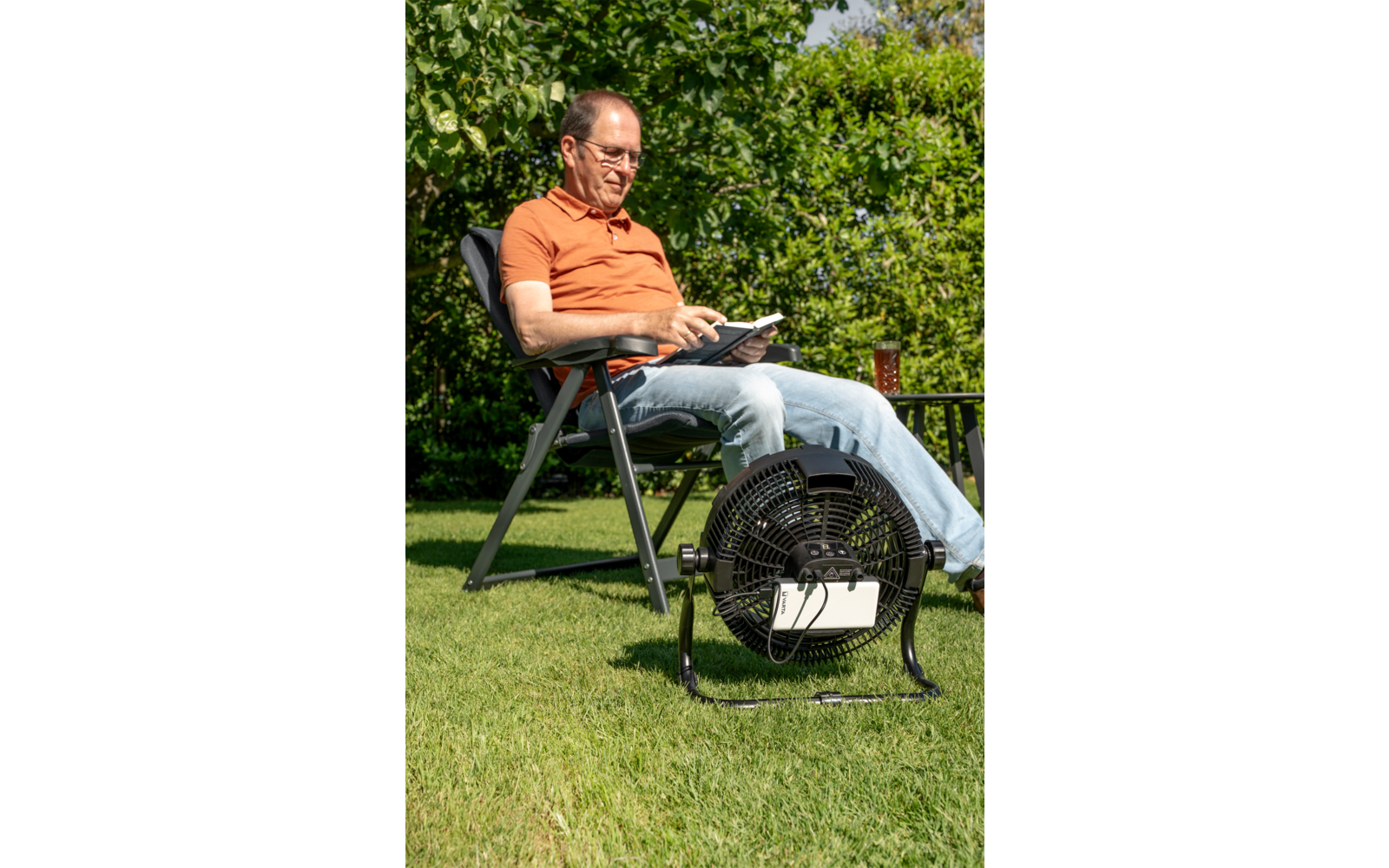 Person sitzt auf Gartenstuhl auf Rasen und hält ein Tablet; tragbarer Ventilator steht davor, Beistelltisch mit Getränk, Hecken im Hintergrund