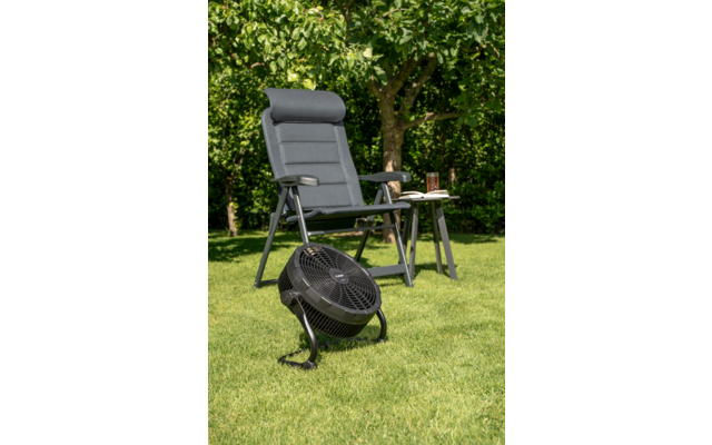 Gepolsterter grauer Gartenstuhl neben kleinem Beistelltisch mit offenem Buch und Glas. Schwarzer tragbarer Ventilator auf dem Rasen. Hecke und Baum im Hintergrund