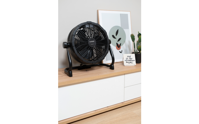 Schwarzer Tischventilator auf einer Holzkommode, daneben ein gerahmter abstrakter Druck, zwei kleine Kakteentöpfe und eine kleine Karte mit Text