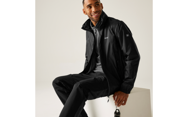 Mann sitzt auf weißem Block, trägt schwarze Jacke, graues Shirt und schwarze Hose, lächelnd, hält Trinkflasche mit Karabiner