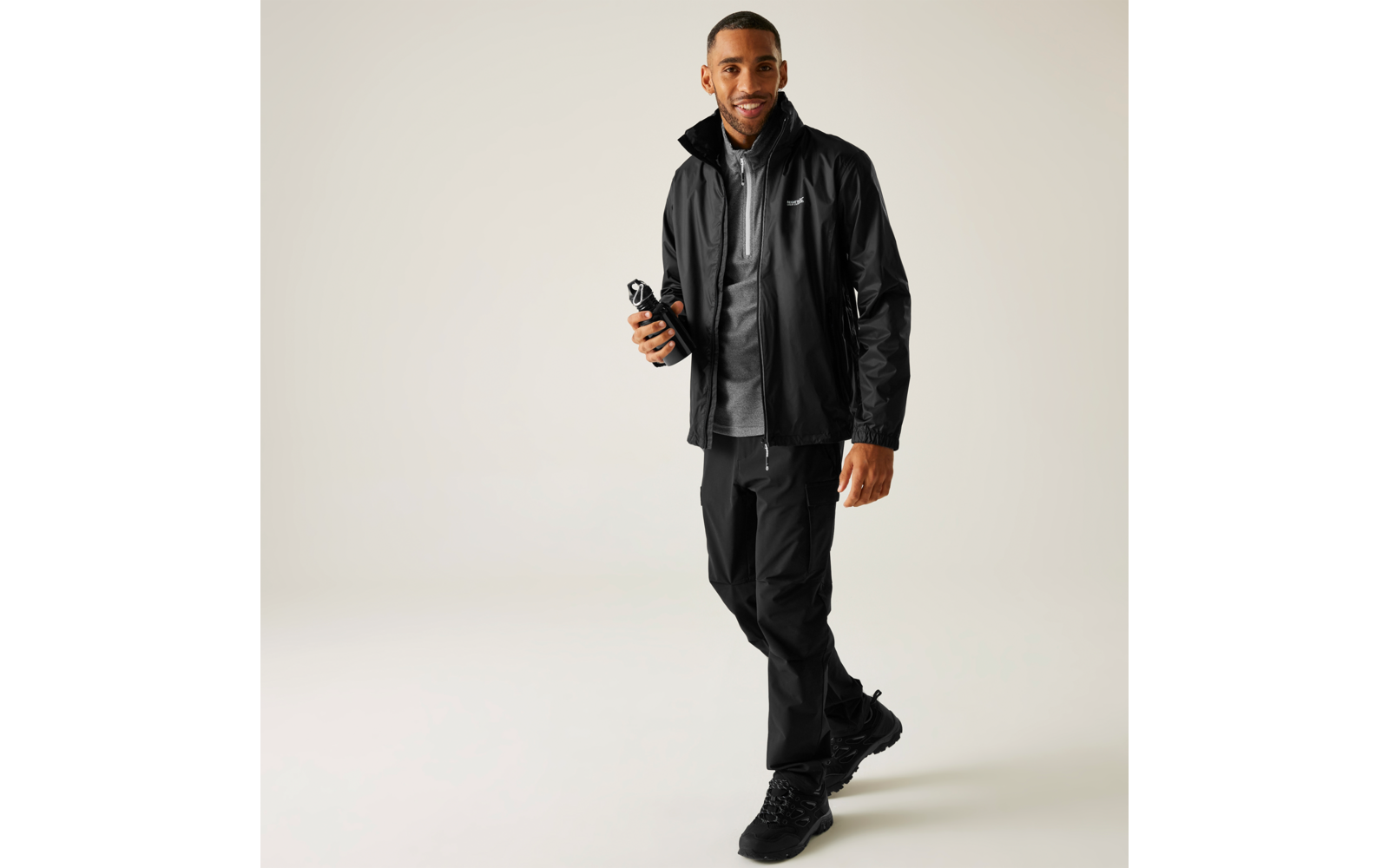 Mann trägt schwarze Regenjacke, graues Oberteil, schwarze Hose und schwarze Schuhe und hält einen zusammengeklappten Regenschirm in der rechten Hand vor hellem Hintergrund