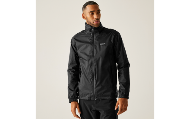 Ein Mann trägt eine schwarze Jacke mit Reißverschluss, kleinem Brustlogo und zwei seitlichen Taschen sowie eine schwarze Hose; er steht vor hellem Hintergrund, schaut nach rechts und hat die Hände an den Seiten