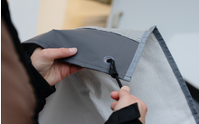 Zwei Hände ziehen eine schwarze Kordel mit Kunststoffstopper durch eine Metallöse in grauem Stoff mit heller Innenlage