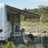 Person sitzt auf Campingstuhl unter ausgefahrener Markise neben Wohnmobil und liest ein Buch; Hund liegt auf dem Gras; Busch- und Baumlandschaft im Hintergrund