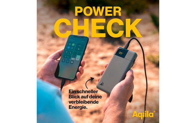 Main tenant un smartphone avec une application ouverte, à côté d'une batterie externe noire avec câble sur un sol sablonneux