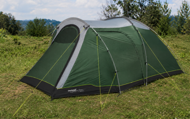 Groene, langwerpige tent met grijs dak en gele scheerlijnen op gras; bomen en bewolkte lucht op de achtergrond