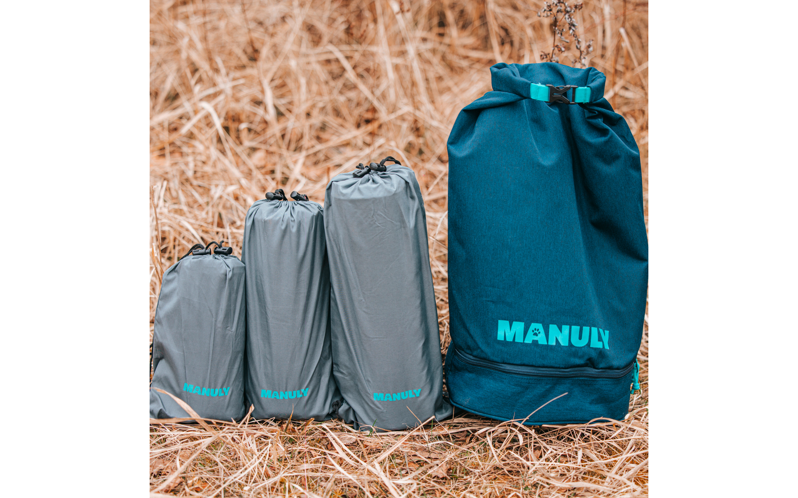 Drei graue, gepackte Packbeutel und eine größere türkisblaue Rolltop-Tasche mit der Aufschrift "MANULY" auf trockenem Gras