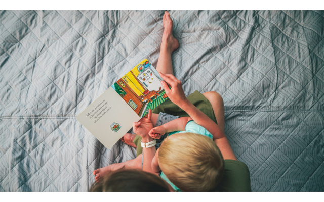 Vogelperspektive auf zwei Personen auf einer grauen Steppbettdecke; eine größere Person hält ein geöffnetes buntes Kinderbuch, eine kleinere Person sitzt daneben; Hände und Füße sichtbar