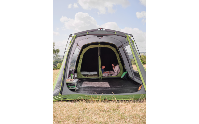 Offenes grünes Campingzelt auf einer Wiese; im Inneren sitzen zwei Personen auf Schlafunterlagen; kleiner Beistelltisch links; Himmel mit Wolken