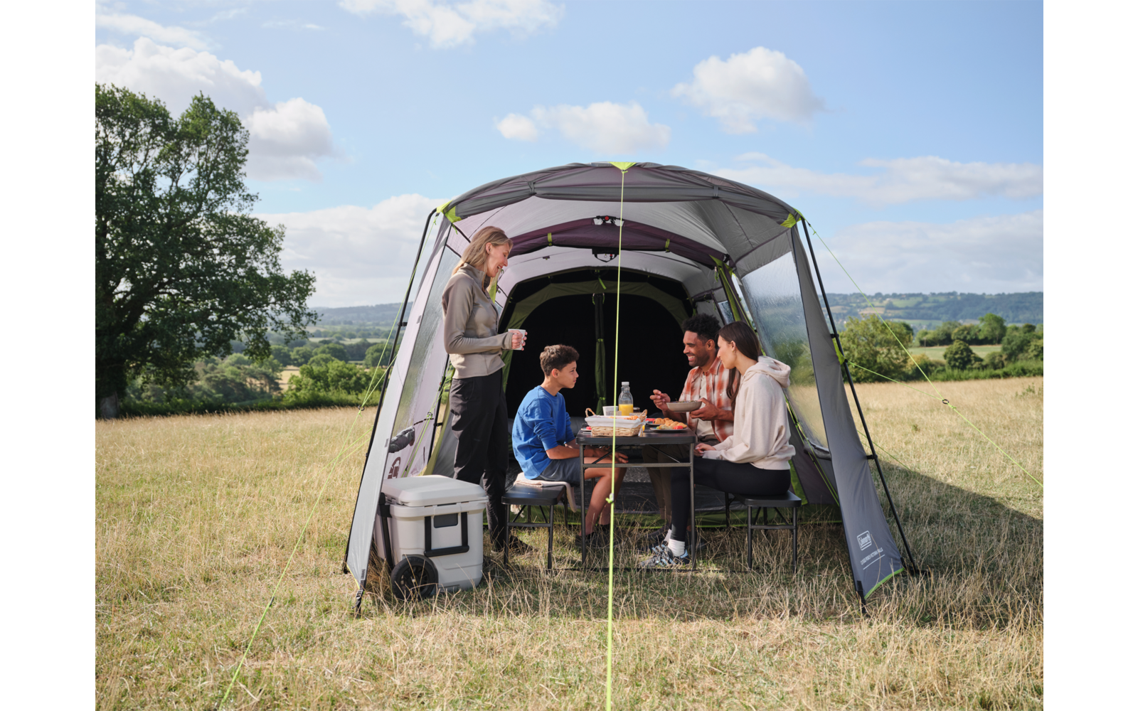 Vier Personen sitzen und stehen in einem offenen Campingzelt um einen Klapptisch mit Geschirr; links vorne eine Kühlbox, im Hintergrund Wiese, ein Baum und bewölkter Himmel