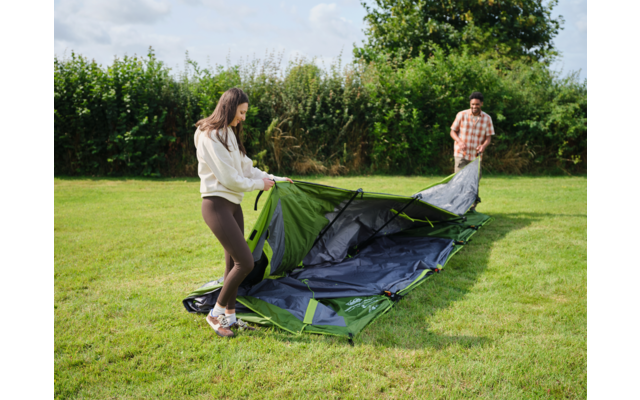Zwei Personen auf einer Wiese neben einem teilweise aufgebauten grünen Zelt; eine Person hält die Zeltplane, die andere steht im Hintergrund
