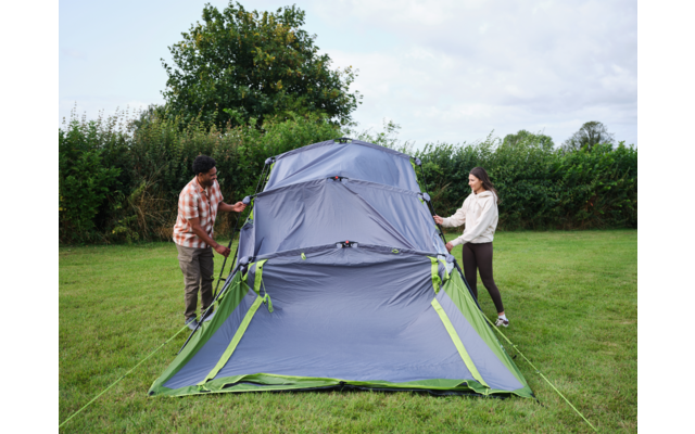 Zwei Personen halten die Seiten eines aufgestellten blau-grauen Zelts mit grünen Akzenten auf einer Wiese; Hecke und Bäume im Hintergrund, bewölkter Himmel