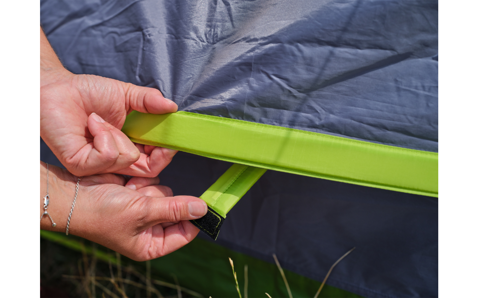 Zwei Hände halten einen grünen Gurt mit Klett vor einem grauen Stoff; Gras am unteren Bildrand; Armbänder an den Handgelenken