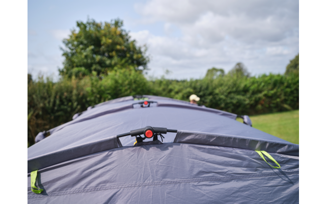 Mehrere grau-blaue Zelte in einer Reihe auf einer Wiese, im Vordergrund ein roter Verschluss an einem Zelt, im Hintergrund Büsche und bewölkter Himmel