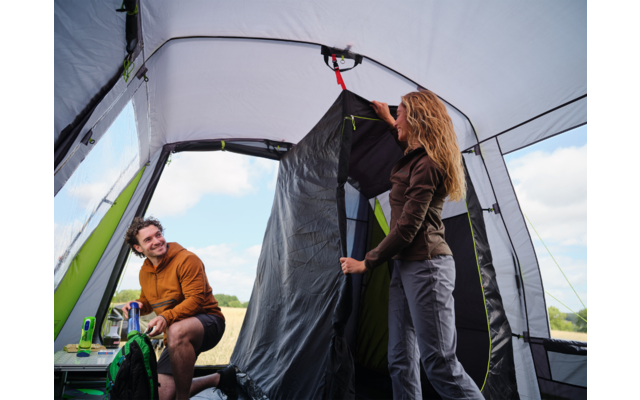 Zwei Personen in einem Zelt: eine Person mit langen blonden Haaren steht und öffnet eine Innentrennwand, eine sitzende Person in oranger Jacke lächelt; Campingstühle, Rucksack und Felder durch Zeltfenster sichtbar