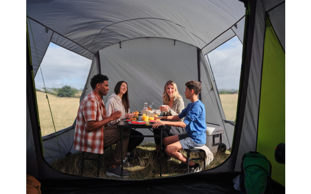 Vier Personen sitzen an einem Tisch in einem offenen Campingzelt, auf dem Tisch stehen Teller, Becher und Essen, im Hintergrund eine Wiese