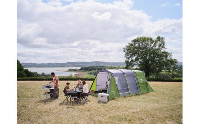 Großes grünes Zelt auf einer Wiese; mehrere Personen sitzen an einem Campingtisch, eine Person steht an einem Campingkocher; Kühlbox in der Nähe, See im Hintergrund, Baum und bewölkter Himmel