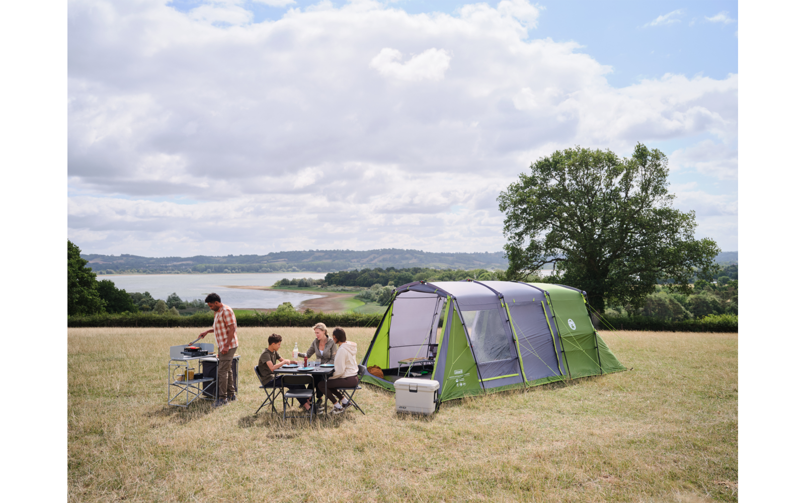 Großes grünes Zelt auf einer Wiese; mehrere Personen sitzen an einem Campingtisch, eine Person steht an einem Campingkocher; Kühlbox in der Nähe, See im Hintergrund, Baum und bewölkter Himmel