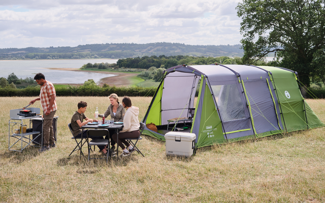Tienda grande y verde en un campo de hierba; varias personas sentadas en una mesa de camping, una persona de pie junto a una cocina portátil; nevera portátil cerca, lago al fondo, un árbol y cielo nublado