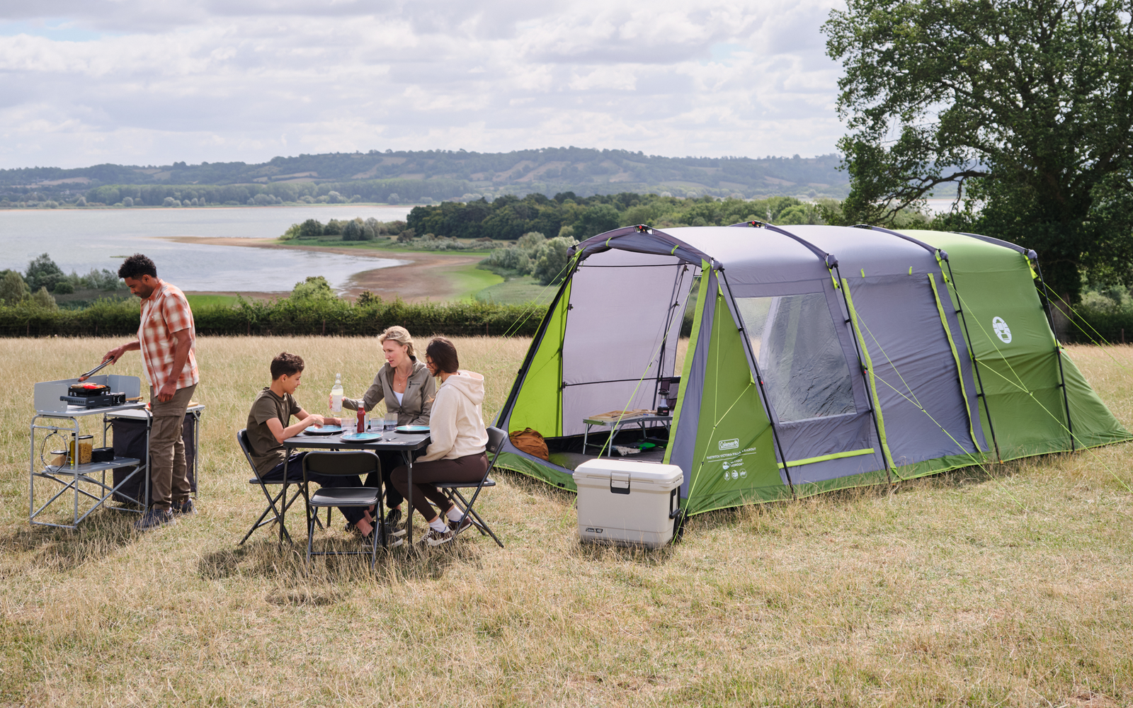 Tienda grande y verde en un campo de hierba; varias personas sentadas en una mesa de camping, una persona de pie junto a una cocina portátil; nevera portátil cerca, lago al fondo, un árbol y cielo nublado