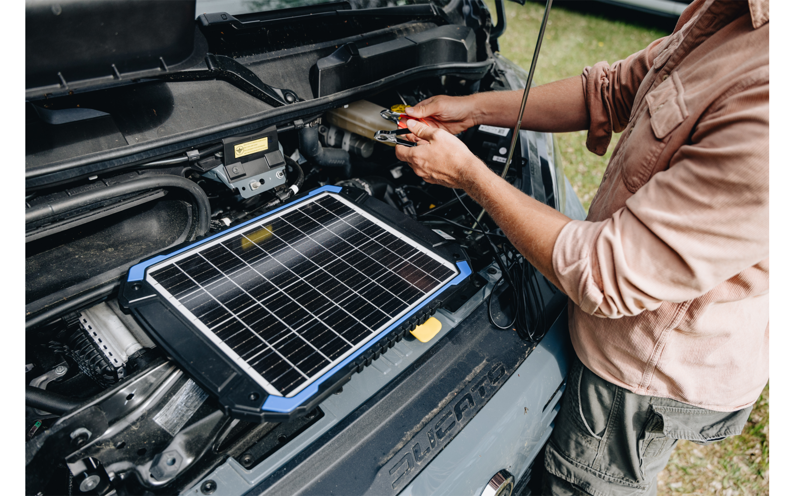 Persona tiene pinze rosse e nere sui morsetti della batteria accanto a un motore aperto; un pannello solare portatile è appoggiato sul motore