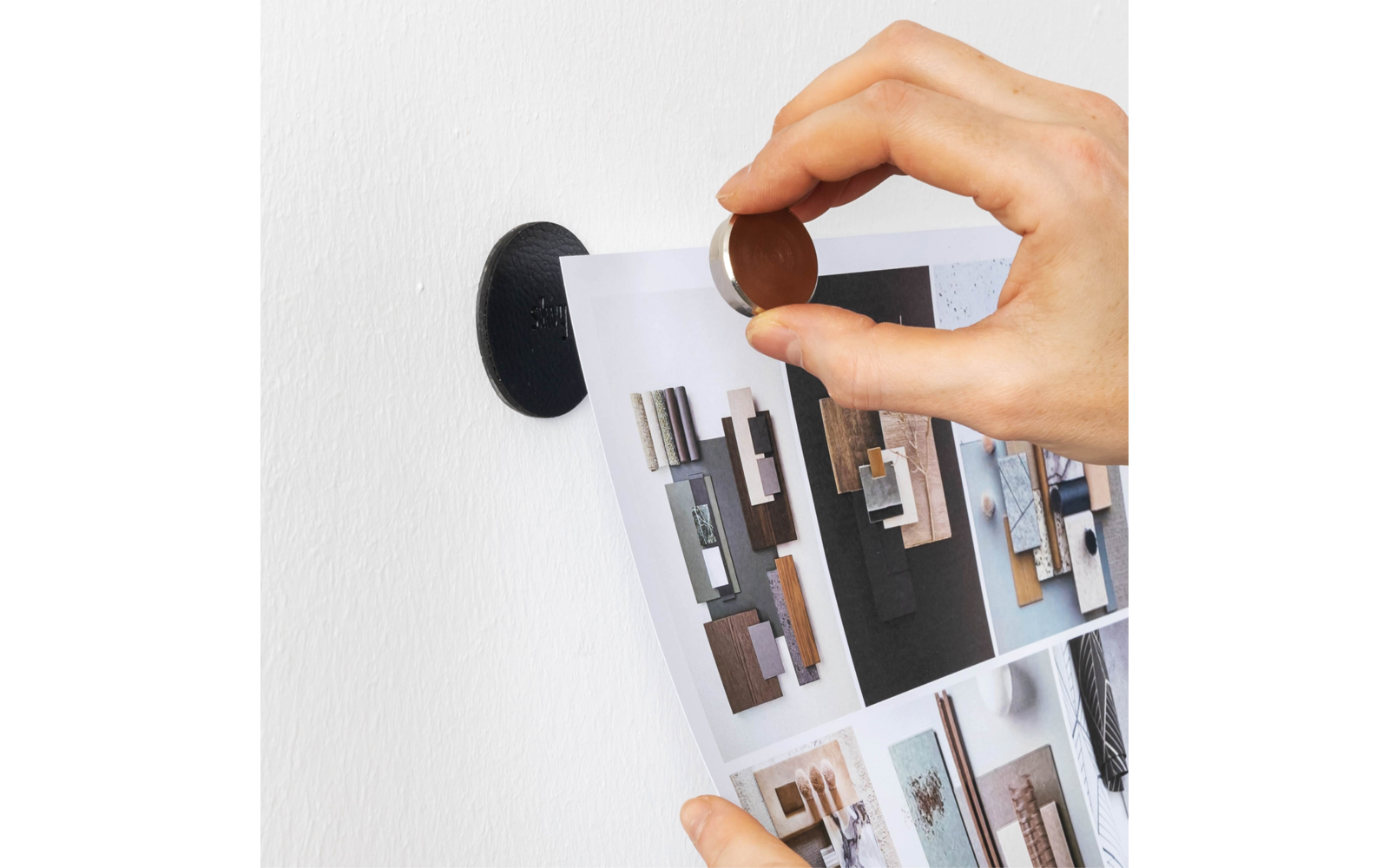 Hand hält eine Münze gegen einen runden schwarzen Magnet an einer weißen Wand; die Münze hält ein bedrucktes Blatt mit kleinen Fotos von Innenelementen