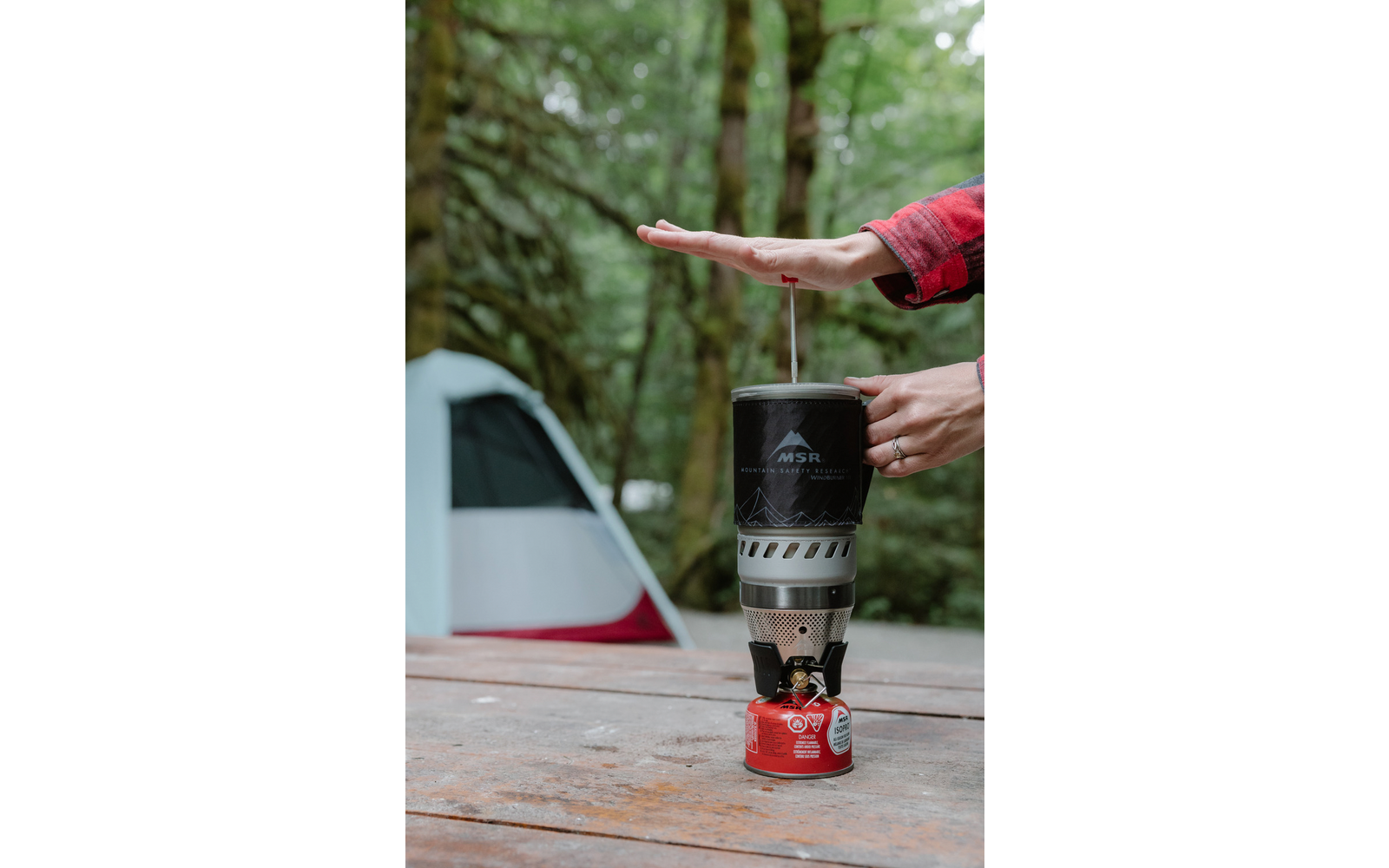 Fornello a gas con bomboletta rossa su tavolo di legno; recipiente da cucina argentato; una mano sopra il recipiente, una mano che tiene la maniglia; tenda e bosco sullo sfondo