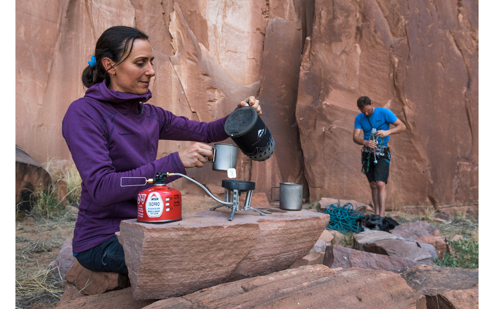 Persona con giacca viola versa acqua da un bollitore in una tazza su un fornello da campeggio appoggiato su una roccia; sullo sfondo un'altra persona con T‑shirt blu sistema corde da arrampicata vicino a una parete di arenaria