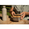 Hands of a person holding a smartphone above a stainless steel lunchbox with a bamboo lid, a beige water bottle to the left on a wooden table, blurred background