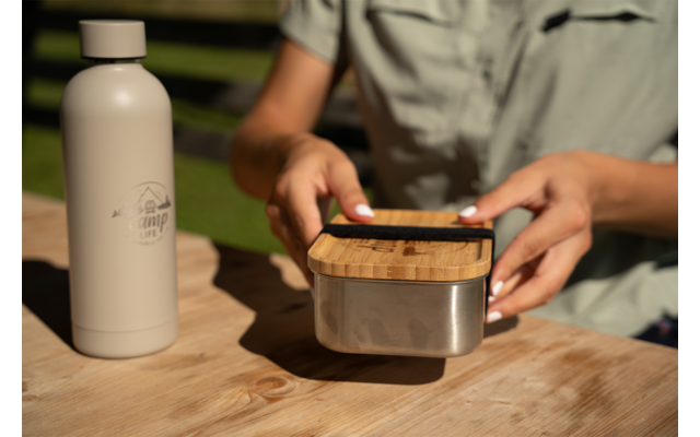 Hands of a person holding a smartphone above a stainless steel lunchbox with a bamboo lid, a beige water bottle to the left on a wooden table, blurred background