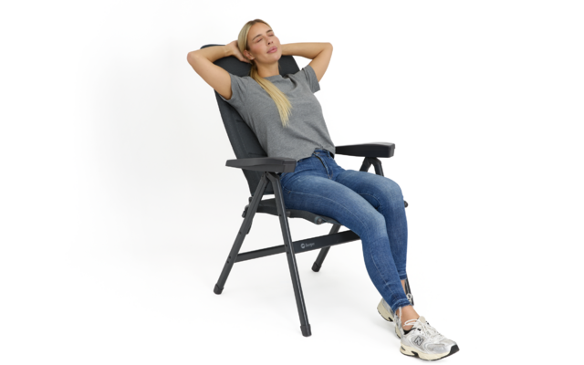 Frau sitzt in klappbarem Liegestuhl, lehnt sich zurück mit Händen hinter dem Kopf, trägt graues T-Shirt, blaue Jeans und Turnschuhe, weißer Hintergrund